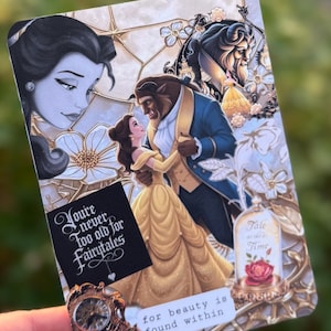 May include: A rectangular card featuring a collage of Beauty and the Beast characters, including Belle and the Beast dancing. The card displays the text "You're never too old for Fairytales" and "for beauty is found within," alongside a rose in a glass dome.