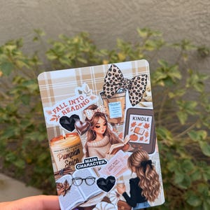 May include: A rectangular sticker featuring a collage of autumn-themed illustrations including a pumpkin spice candle, an open book, a Kindle e-reader, a coffee cup with a leopard print bow, and text reading Fall Into Reading and Main Character.