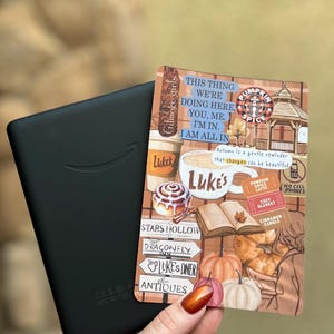 May include: A black e-reader device next to a rectangular sticker featuring autumn-themed illustrations, including a Luke's Diner coffee cup, a cinnamon roll, an open book, pumpkins, and text that reads Gilmore Girls, This thing we're doing here, you, me. I'm in. I am all in, and Stars Hollow.