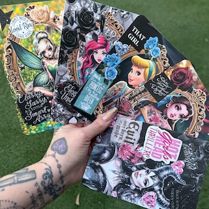 May include: A hand holds several decorative cards featuring fairy tale characters and gothic designs. The cards have text like "Classy, Sassy, & a Little Smart Assy" and "I'm not a Princess, I'm the fucking Queen."