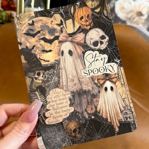 May include: A rectangular card featuring a collage of Halloween imagery including two ghosts with bows, a skeleton in a witch hat, skulls, a full moon, bats, and spiderwebs. The text reads Stay SPOOKY and May the only Spirits that invade your home at Halloween be the happy Spirits of friends and Book of Shadows.