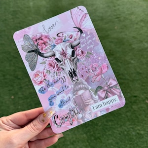 May include: A pink rectangular card with a floral and skull design. The card features a cow skull, roses, butterflies, and the words "love", "Be the energy you want to attract", "Cowgirl", "Hey you, don't give up okay?", and "I am happy."