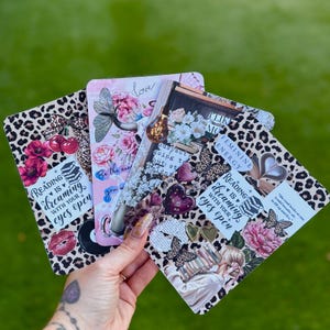 May include: A hand holding four decorative cards with various designs. The cards feature floral, butterfly, and leopard print patterns. Text includes "Reading is dreaming with your eyes open" and "Love".