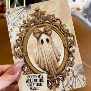 May include: A rectangular card featuring a central illustration of a ghost wearing a brown bow, framed by an ornate gold oval border. The background includes spiderweb and floral patterns. A white label at the bottom reads, "BOOKS MAY WELL BE THE ONLY TRUE MAGIC."