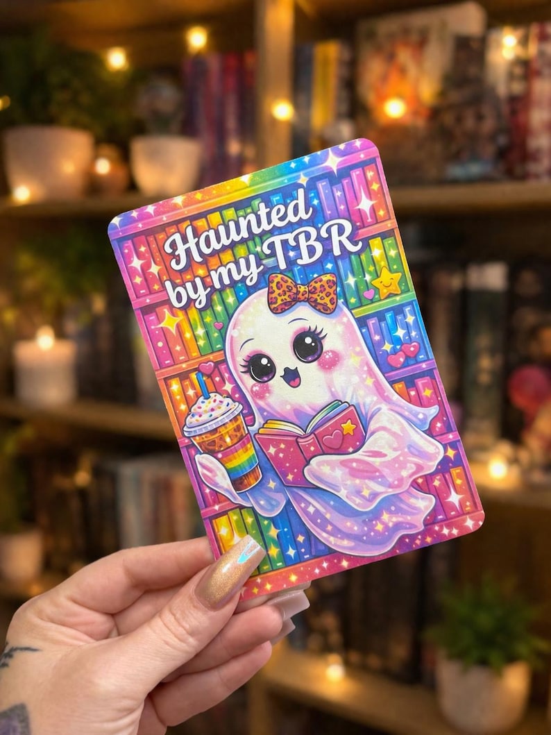 May include: A colorful rectangular sticker with the text "Haunted by my TBR." The sticker features a cute ghost character with a bow, holding a book and a rainbow-colored drink. The background is a rainbow bookshelf.
