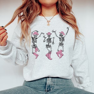 Puede incluir: Una sudadera gris con un gráfico de tres esqueletos con sombreros de vaquero y botas rosas. Los esqueletos están bailando.