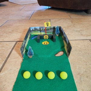 May include: A miniature golf course with a green putting surface, a cardboard cutout of a golf course, and four yellow golf balls. The course has a small wooden bridge and a flag with a smiley face.