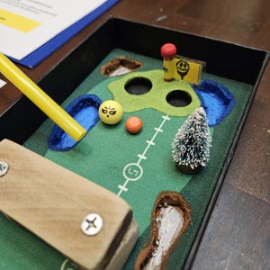 May include: Miniature golf course diorama with a green felt playing surface. Includes a wooden putter, yellow golf ball with a face, orange golf ball, and a small Christmas tree. A yellow flag with a smiley face is also present.