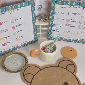 Può includere: Una collezione di oggetti decorativi, tra cui un sottobicchiere a forma di orso, una tazza con manico arancione e un piatto decorativo. Lo sfondo presenta una carta da regalo a tema Hello Kitty e due lavagne con elenchi di articoli.