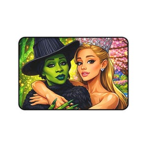 Op de afbeelding: Een rechthoekige muismat met een kleurrijke illustratie van twee vrouwen. De ene is een groenhuidige heks met een zwarte hoed en de andere een blonde vrouw met een kroon. De achtergrond toont een bos en roze bloesem bomen.