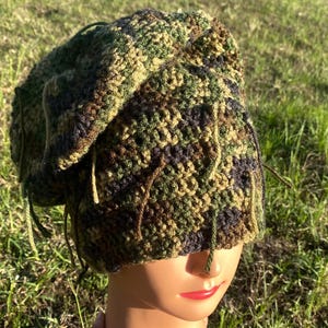 May include: A camouflage-patterned crochet beanie hat in shades of green, brown, and black. The hat has a slouchy fit and features dangling yarn strands. The hat is displayed on a mannequin head.