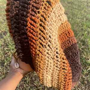 Könnte beinhalten: Ein handgehäkelter Hut in Braun-, Beige- und Orangetönen. Der Hut hat ein gestreiftes Design mit einer offenen, strukturierten Webart. Ein kleiner Muschelanhänger hängt an der Seite. Der Hut wird vor einem Hintergrund aus grünem Gras gehalten.