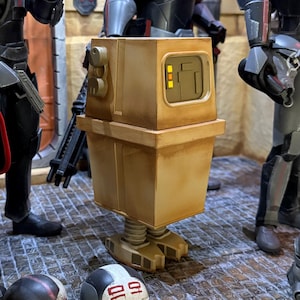 Puede incluir: Cinco figuras de acción, incluyendo un droide, están posadas en una escena. Las figuras están vestidas con armadura negra y roja y sostienen armas. El droide es un pequeño robot en forma de caja con un cuerpo amarillo y un panel negro en la parte delantera.