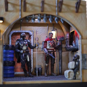 May include: A miniature diorama featuring two action figures in a desert-themed setting. The figures are dressed in futuristic armor and clothing. The diorama includes a variety of details, such as a door, a barrel, and a wall with a window.