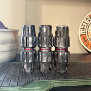 May include: Three metallic gray connectors with a red band, arranged vertically. The connectors have a ribbed design and are likely part of a larger set or system. The objects are displayed on a textured surface.