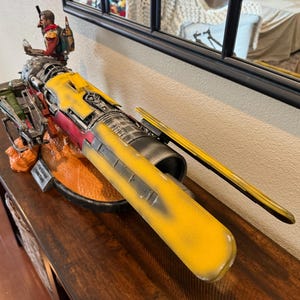 Puede incluir: Un modelo detallado de una nave espacial amarilla, roja y gris con una figura de una persona de pie en la parte superior. El modelo se exhibe sobre una superficie de madera. La nave espacial tiene un diseño largo y elegante con detalles intrincados.