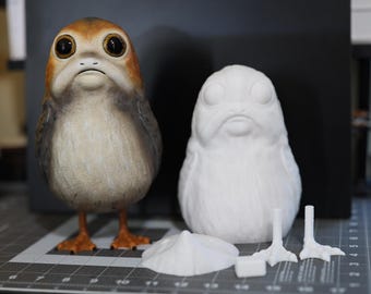 May include: A painted figurine of a Porg from Star Wars: The Last Jedi. The figurine is brown and white with large eyes. A white unpainted version of the figurine is also shown, along with separate unpainted feet.