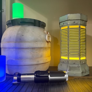 May include: A collection of sci-fi themed decorative items. Includes a cylindrical light with yellow illumination, a large, weathered container with a green light on top, and a silver and black lightsaber prop.
