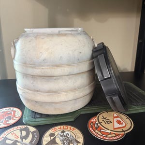 May include: A weathered, off-white cylindrical container with a removable dark gray lid. The container has horizontal bands and decorative elements. Several circular stickers with various designs and text are visible.