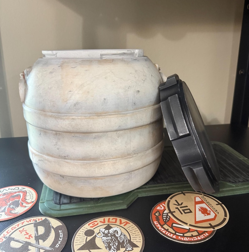 May include: A weathered, off-white cylindrical container with a removable dark gray lid. The container has horizontal bands and decorative elements. Several circular stickers with various designs and text are visible.
