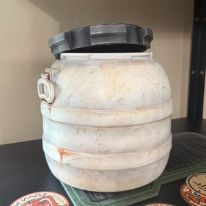 May include: A weathered, cylindrical container with a removable, dark gray lid. The container has horizontal bands and a handle on the side. The exterior is off-white with brown staining. Several circular stickers are visible near the base.