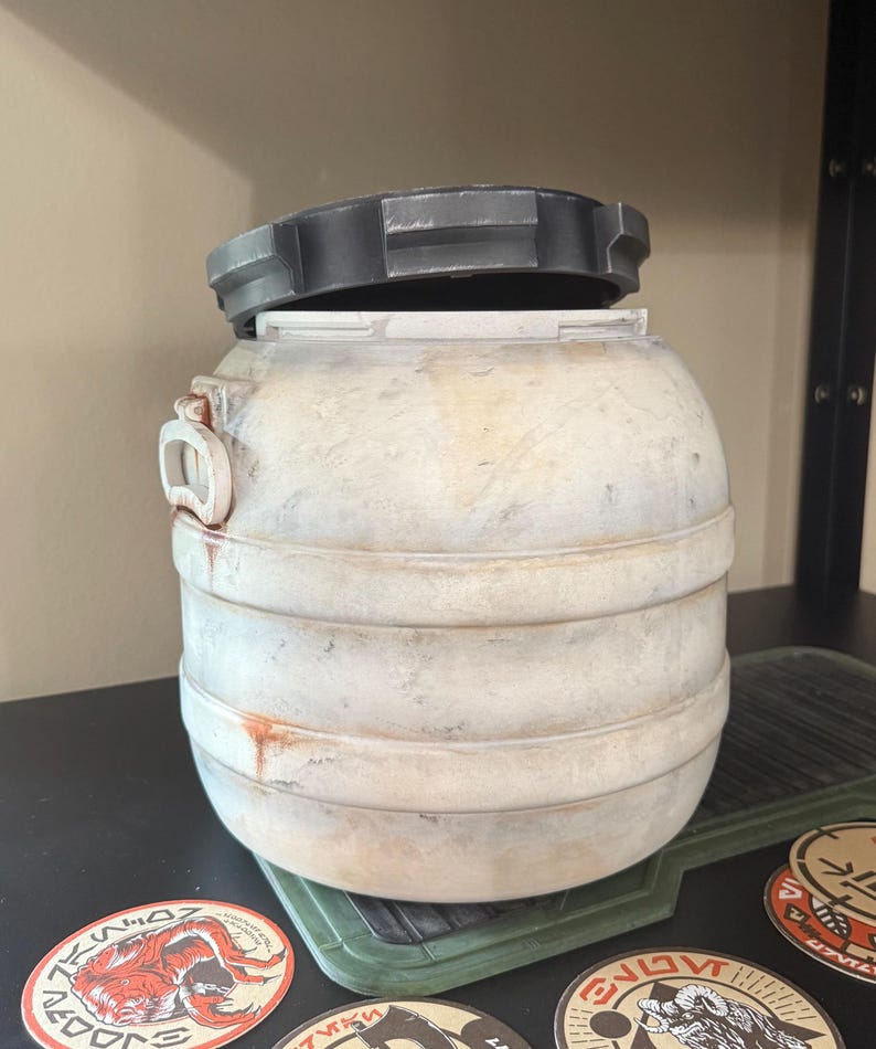 May include: A weathered, cylindrical container with a removable, dark gray lid. The container has horizontal bands and a handle on the side. The exterior is off-white with brown staining. Several circular stickers are visible near the base.