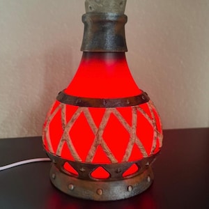 May include: A decorative bottle with a red glowing interior, featuring a textured exterior with a crisscross pattern. The bottle has a dark, metallic-looking base and a cork stopper. The overall design suggests a vintage or fantasy theme.