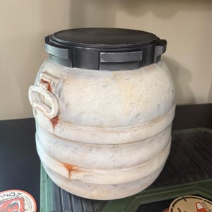 May include: A cylindrical, off-white container with a black, rectangular lid. The container has a weathered appearance with horizontal bands and a handle-like feature on the side. The design suggests a vintage or industrial aesthetic.