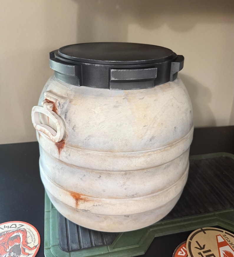 May include: A cylindrical, off-white container with a black, rectangular lid. The container has a weathered appearance with horizontal bands and a handle-like feature on the side. The design suggests a vintage or industrial aesthetic.