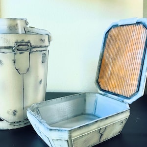 May include: A white, weathered, rectangular box with a hinged lid. The lid is open, revealing a brown, textured interior. The box is made of a hard, durable material and has a distressed look.