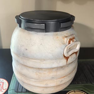 May include: A weathered, cylindrical container with a black lid and handle. The container has a light beige finish with dark accents and a textured surface. The lid has a rectangular design. The container is sitting on a green surface.