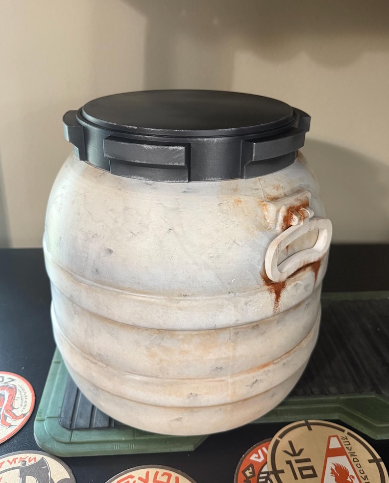 May include: A weathered, cylindrical container with a black lid and handle. The container has a light beige finish with dark accents and a textured surface. The lid has a rectangular design. The container is sitting on a green surface.