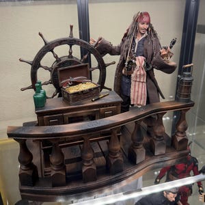 Può includere: Diorama dettagliato con una scena di nave pirata. La scena comprende un timone, uno scrigno del tesoro pieno d'oro e una figura di pirata. La tavolozza dei colori è dominata da marroni, ori e verdi, creando una miniatura realistica e dettagliata.