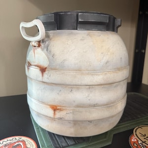 May include: A weathered, cylindrical container with a black top and a white handle. The body is off-white with horizontal bands and rust-colored streaks. The container sits on a green, textured surface, with several circular stickers visible.