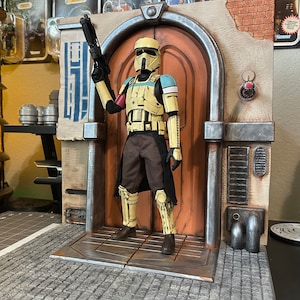 May include: A yellow and blue Star Wars action figure stands in front of a doorway with a brown door. The figure is holding a blaster rifle in its right hand. The figure is standing on a platform with a brick pattern.