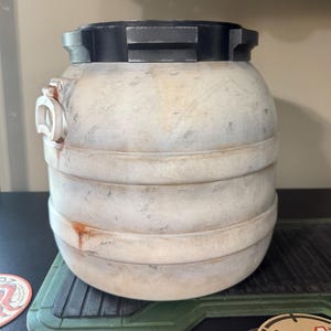 May include: A cylindrical, off-white container with horizontal ridges and a black, rectangular top. The container has a weathered appearance with rust-colored accents and a small, white, curved handle on the side. It sits on a green, textured surface.