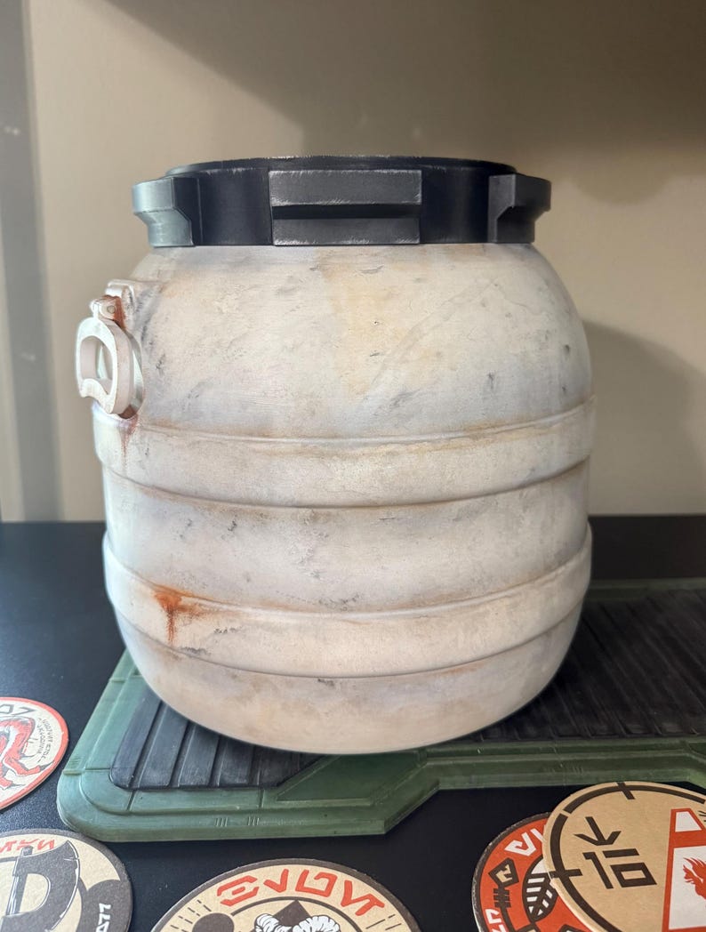 May include: A cylindrical, off-white container with horizontal ridges and a black, rectangular top. The container has a weathered appearance with rust-colored accents and a small, white, curved handle on the side. It sits on a green, textured surface.