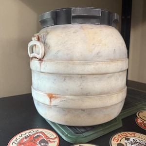 May include: A weathered, off-white barrel with a dark gray lid and accents. The barrel has horizontal bands and a small handle. Several circular stickers with various designs are visible near the base of the barrel.