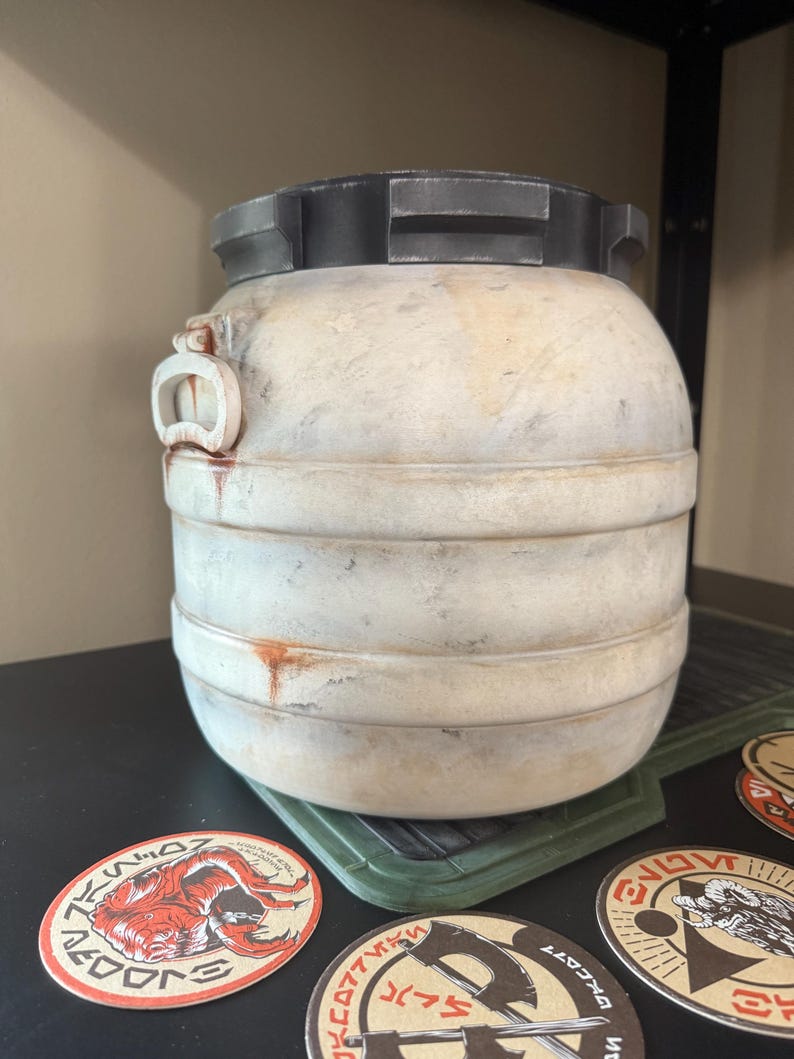 May include: A weathered, off-white barrel with a dark gray lid and accents. The barrel has horizontal bands and a small handle. Several circular stickers with various designs are visible near the base of the barrel.