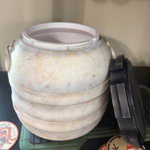 May include: A weathered, off-white barrel with a removable black lid. The barrel has horizontal bands and a textured surface. The lid is partially open, revealing the interior. The design suggests a vintage or rustic aesthetic.