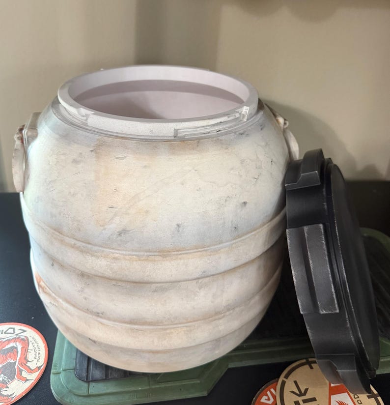 May include: A weathered, off-white barrel with a removable black lid. The barrel has horizontal bands and a textured surface. The lid is partially open, revealing the interior. The design suggests a vintage or rustic aesthetic.