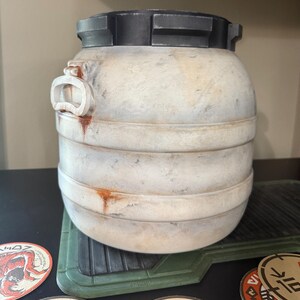 May include: A cylindrical, off-white container with a dark gray lid and accents. The container has horizontal bands and a handle-like feature on the side. It sits on a green, textured surface, with circular coasters visible below.
