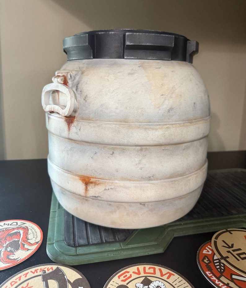 May include: A cylindrical, off-white container with a dark gray lid and accents. The container has horizontal bands and a handle-like feature on the side. It sits on a green, textured surface, with circular coasters visible below.