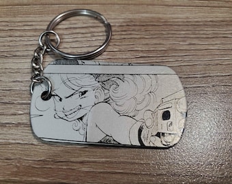 May include: A silver keychain with a black and white image of a woman with a gun. The woman has long hair and is wearing a sleeveless shirt. The keychain is attached to a silver chain.