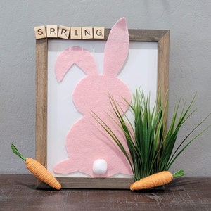 May include: A pink felt bunny silhouette with a white pom-pom tail in a wooden frame with the word "SPRING" spelled out in wooden letters. Green grass and two orange carrots are in the foreground.