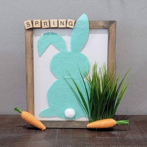 May include: A wooden frame with a light blue felt bunny silhouette and the word "SPRING" spelled out in wooden letters. The frame is decorated with green grass and two orange carrots.