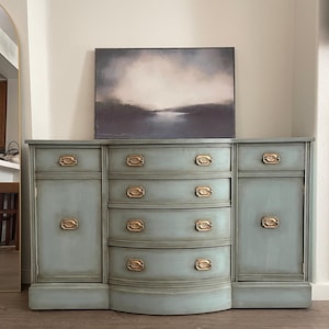 May include: A vintage-style sideboard with a curved front, painted in a light blue-green color. It has three drawers and two cabinet doors with brass hardware. The sideboard is sitting on a wooden floor.