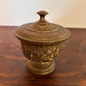 Puede incluir: Un cuenco de latón con tapa que presenta un intrincado grabado floral en el cuerpo y la tapa. El recipiente descansa sobre una base de pedestal circular e incluye un pequeño pomo en la parte superior, con un acabado metálico pulido.