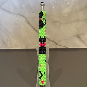 May include: A decorative pen with a vibrant green beaded design. The pen features black and pink accents, with a silver tip and top. The pen is displayed on a clear acrylic stand, showcasing its unique, handcrafted style.