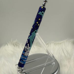 May include: A decorative pen with a silver tip and a barrel covered in blue, white, and yellow rhinestones. The pen is displayed on a clear acrylic stand, set against a white, fluffy background.
